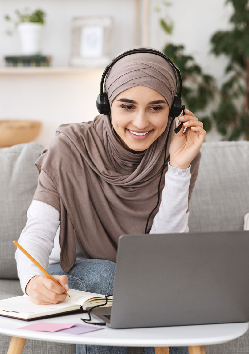 Young woman wearing hijab smiling at laptop while writing notes