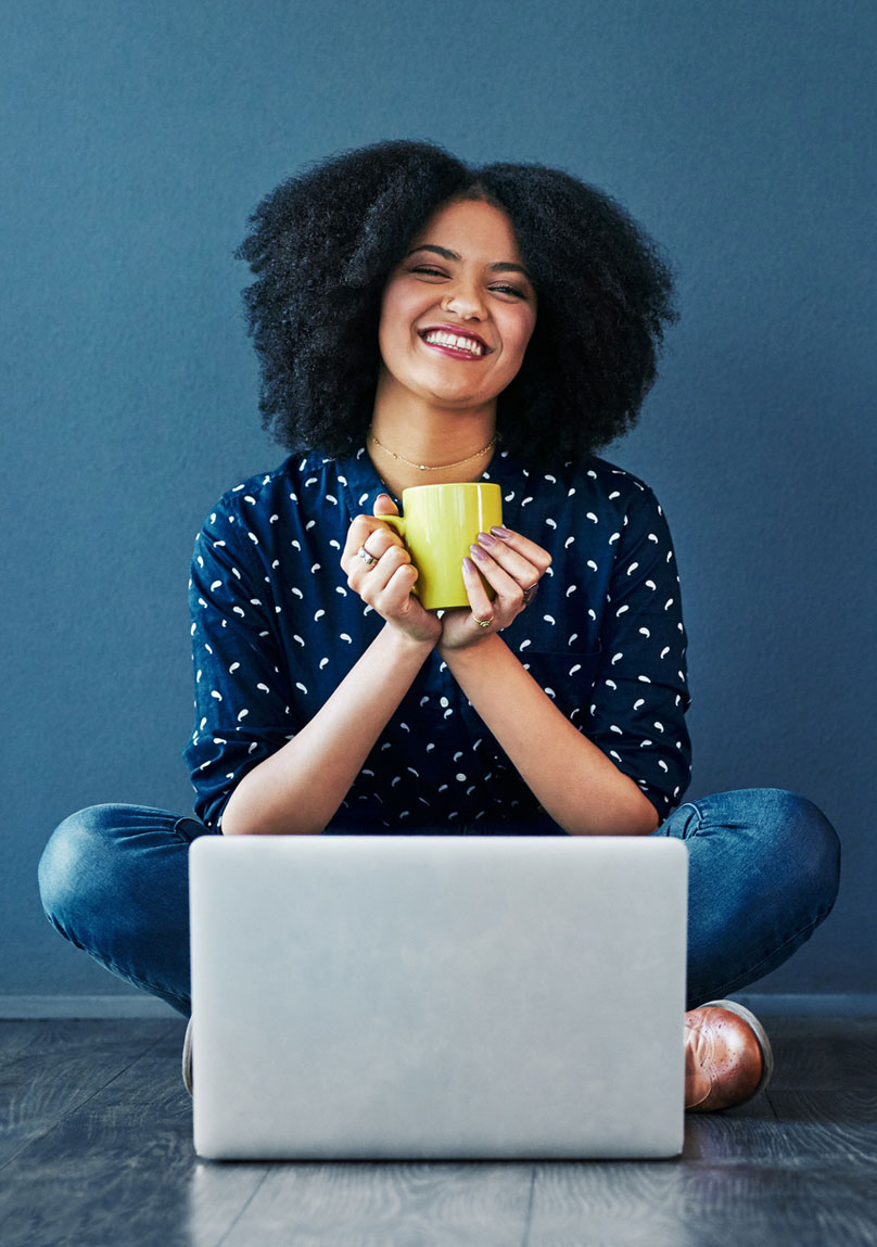 Happy young woman drinking a coffee while sitting cross-legged in front of a laptop
